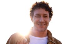 A man with curly hair and a beard smiles warmly at the camera. He is wearing a brown jacket over a white t-shirt. The sun is shining brightly behind him, creating a halo effect around his head. The background is a clear, bright sky.