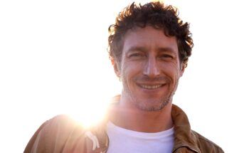 A man with curly hair and a beard smiles warmly at the camera. He is wearing a brown jacket over a white t-shirt. The sun is shining brightly behind him, creating a halo effect around his head. The background is a clear, bright sky.