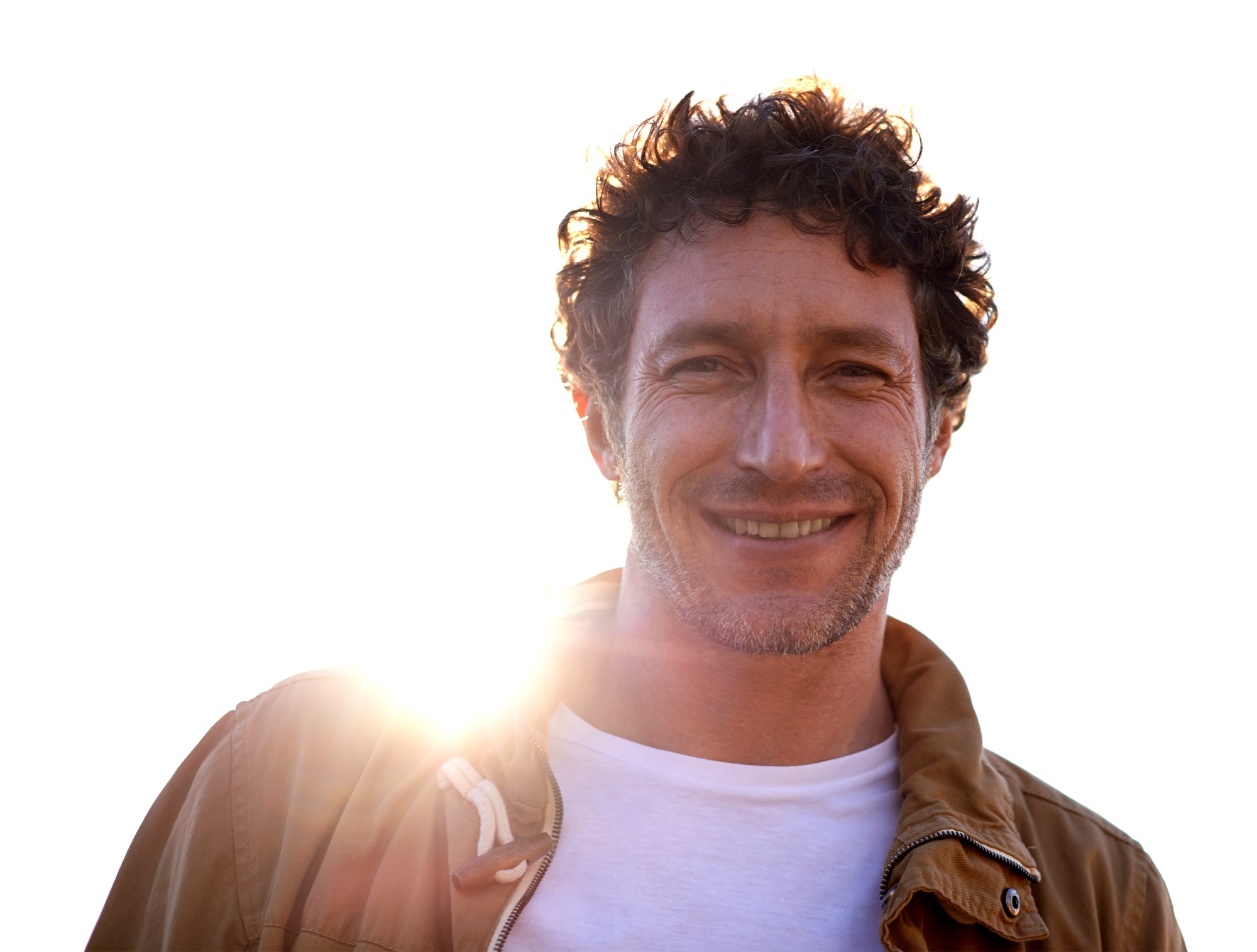 A man with curly hair and a beard smiles warmly at the camera. He is wearing a brown jacket over a white t-shirt. The sun is shining brightly behind him, creating a halo effect around his head. The background is a clear, bright sky.