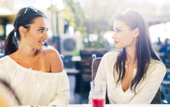 Two women, both with long dark hair, sit at an outdoor café. They are facing each other and smiling. Both are wearing light-colored tops and sunglasses on their heads. A partially filled glass with a red drink is on the table in front of them.