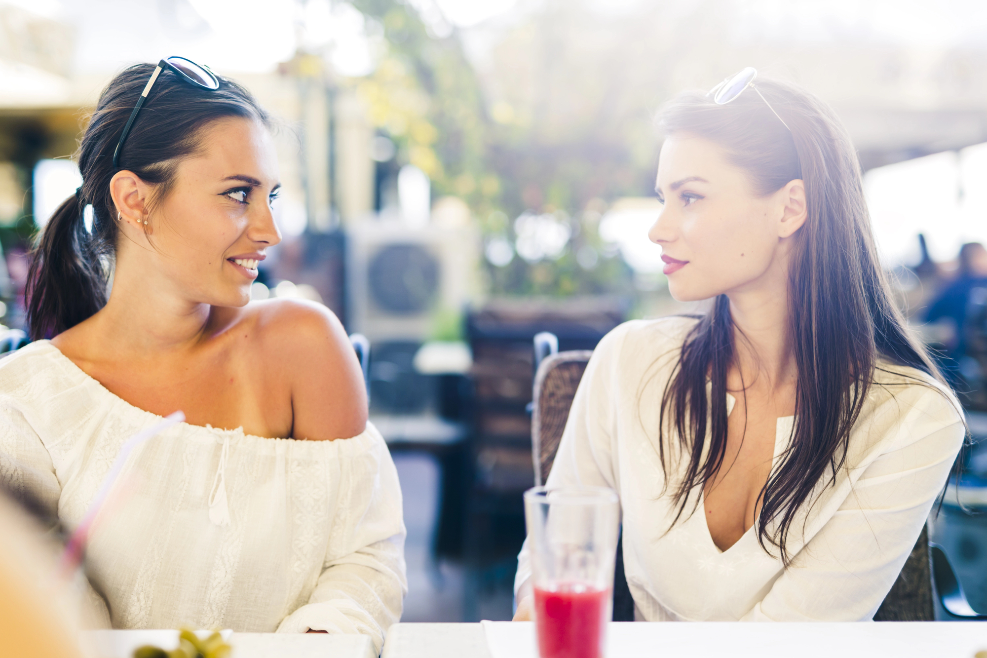 Two women, both with long dark hair, sit at an outdoor café. They are facing each other and smiling. Both are wearing light-colored tops and sunglasses on their heads. A partially filled glass with a red drink is on the table in front of them.