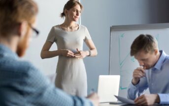 A woman standing and speaking in front of a whiteboard, with two men sitting at a table and listening attentively. One man is holding a pen near his mouth, seemingly deep in thought, while the other is slightly out of focus. The background has soft lighting.