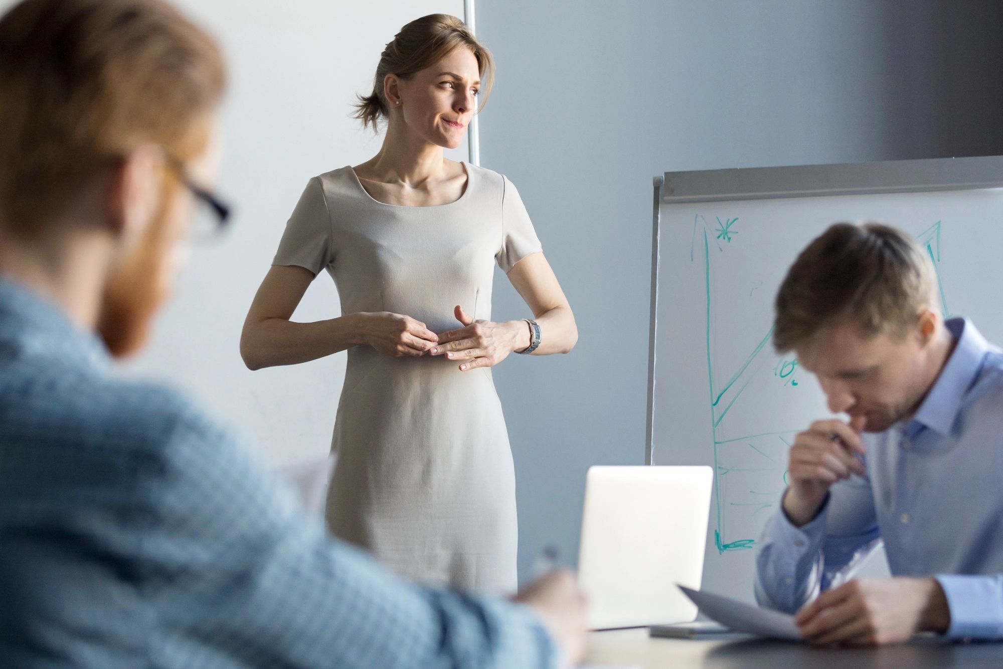 A woman standing and speaking in front of a whiteboard, with two men sitting at a table and listening attentively. One man is holding a pen near his mouth, seemingly deep in thought, while the other is slightly out of focus. The background has soft lighting.