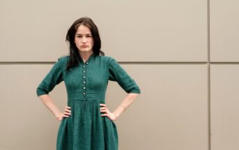 A woman with dark hair stands against a plain beige wall, wearing a green dress with a button-down front. Her hands are on her hips, and she has a serious expression on her face.