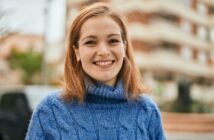 A young woman with shoulder-length brown hair and a blue sweater smiles at the camera while standing outdoors. The background is slightly out of focus, showing a multi-story building and some greenery.