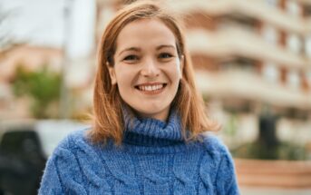 A young woman with shoulder-length brown hair and a blue sweater smiles at the camera while standing outdoors. The background is slightly out of focus, showing a multi-story building and some greenery.