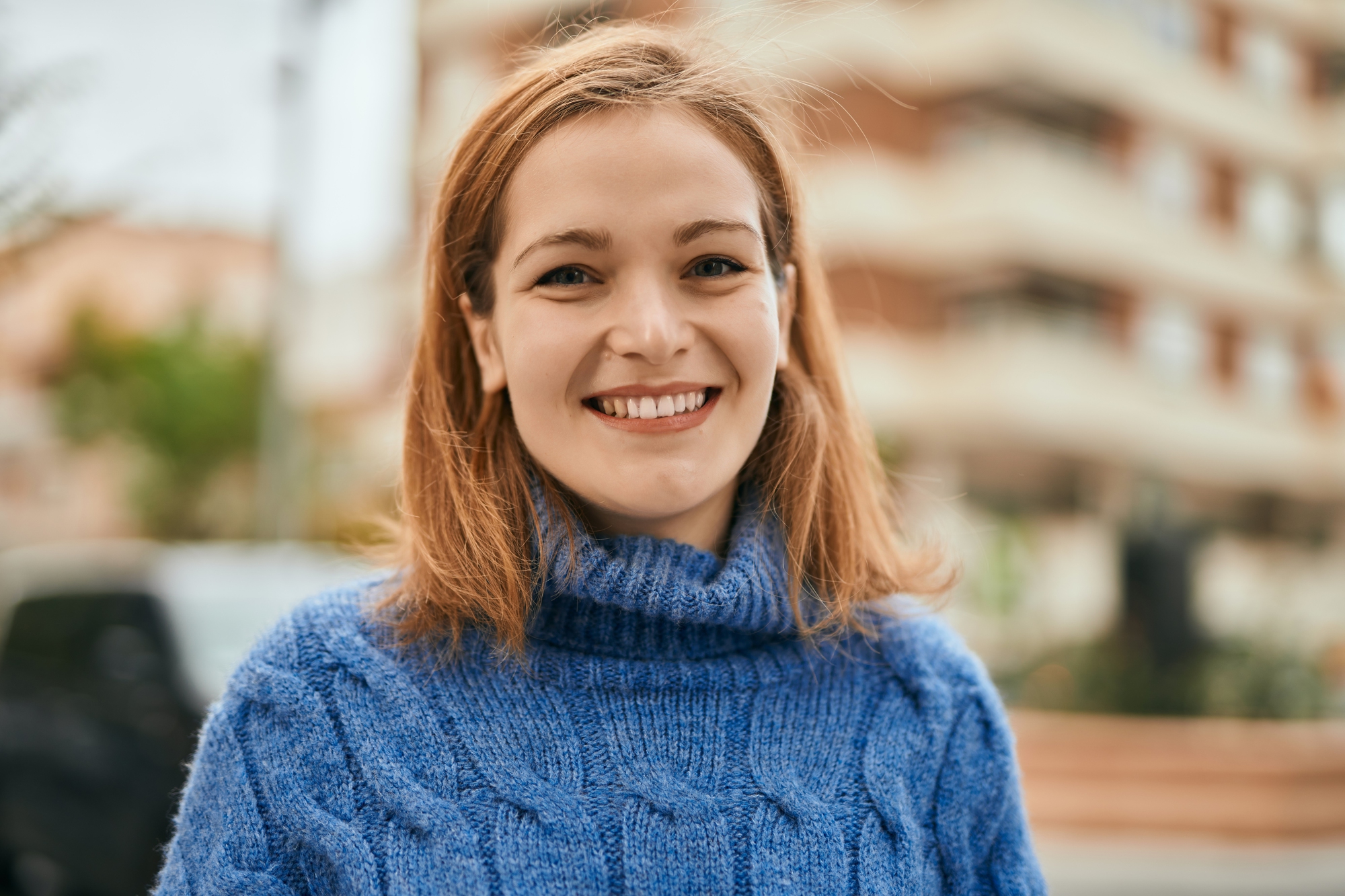 A young woman with shoulder-length brown hair and a blue sweater smiles at the camera while standing outdoors. The background is slightly out of focus, showing a multi-story building and some greenery.