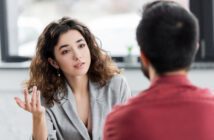 A woman with wavy hair wearing a grey blazer is gesturing with one hand while looking at a man with short dark hair and a red shirt. They appear to be engaged in a conversation in a well-lit room with a blurred background.