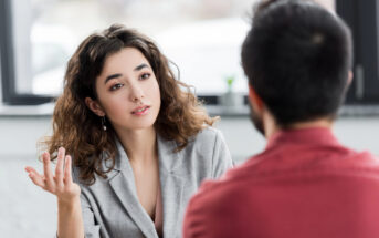 A woman with wavy hair wearing a grey blazer is gesturing with one hand while looking at a man with short dark hair and a red shirt. They appear to be engaged in a conversation in a well-lit room with a blurred background.