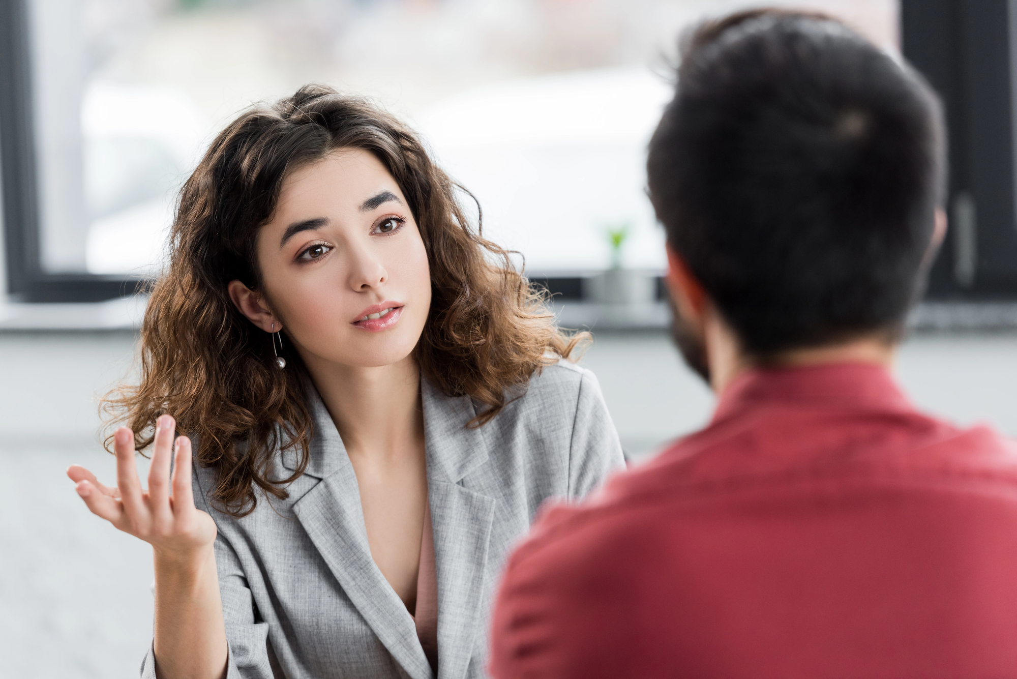 A woman with wavy hair wearing a grey blazer is gesturing with one hand while looking at a man with short dark hair and a red shirt. They appear to be engaged in a conversation in a well-lit room with a blurred background.