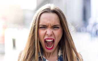 A woman with long blonde hair and red lipstick is standing outdoors and screaming. She is wearing a denim jacket and a white top. The background is blurred, possibly an urban setting.