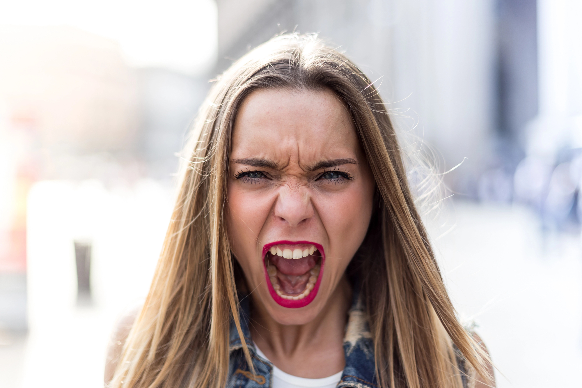 A woman with long blonde hair and red lipstick is standing outdoors and screaming. She is wearing a denim jacket and a white top. The background is blurred, possibly an urban setting.