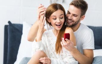 A surprised woman in a white sweater admires an engagement ring held by a smiling man in a white t-shirt sitting behind her. They are indoors, sitting on a bed or couch, with joyful expressions on their faces.