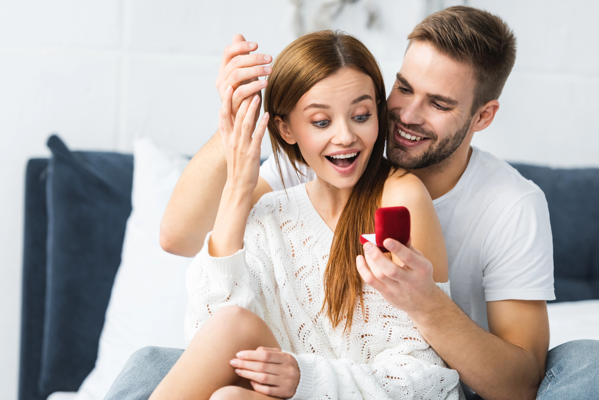 A surprised woman in a white sweater admires an engagement ring held by a smiling man in a white t-shirt sitting behind her. They are indoors, sitting on a bed or couch, with joyful expressions on their faces.