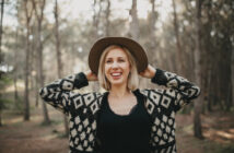 A smiling woman stands in a forest, wearing a black and white patterned sweater, a black top, and a brown felt hat. She has her hands on the hat's brim while looking off into the distance. The background is blurred, showing tall trees with sunlight filtering through.