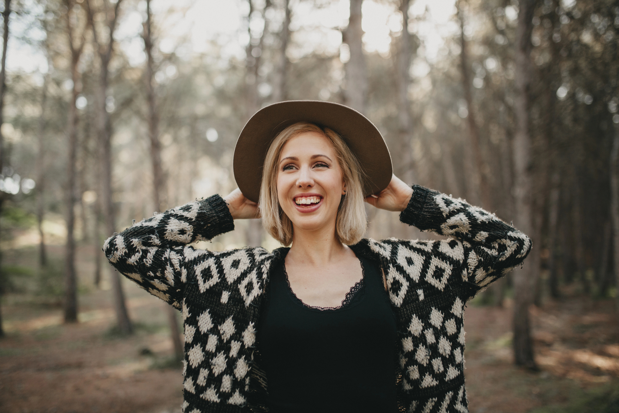 A smiling woman stands in a forest, wearing a black and white patterned sweater, a black top, and a brown felt hat. She has her hands on the hat's brim while looking off into the distance. The background is blurred, showing tall trees with sunlight filtering through.