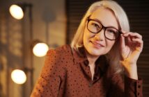 A woman with shoulder-length blonde hair and glasses, wearing a brown polka-dot blouse, smiles and holds her glasses with one hand. She is indoors, with soft lighting and a blurred background showing round lights and shadows.