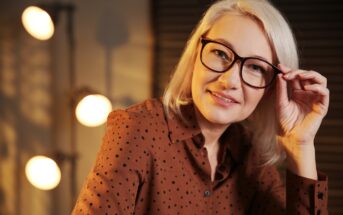 A woman with shoulder-length blonde hair and glasses, wearing a brown polka-dot blouse, smiles and holds her glasses with one hand. She is indoors, with soft lighting and a blurred background showing round lights and shadows.