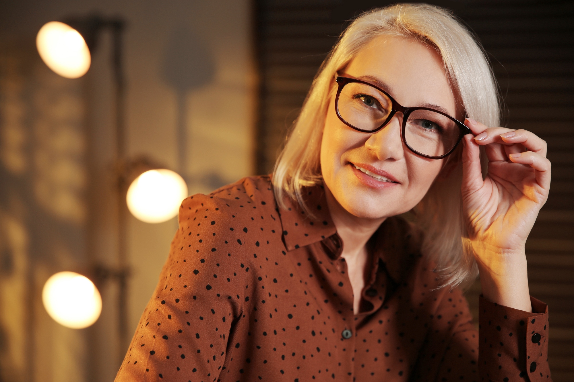 A woman with shoulder-length blonde hair and glasses, wearing a brown polka-dot blouse, smiles and holds her glasses with one hand. She is indoors, with soft lighting and a blurred background showing round lights and shadows.