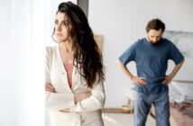 A woman with long dark hair stands by a window with her arms crossed, looking pensive and away from a man in the background who has his hands on his hips, appearing frustrated. The setting looks like a bedroom with soft lighting.