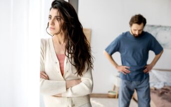 A woman with long dark hair stands by a window with her arms crossed, looking pensive and away from a man in the background who has his hands on his hips, appearing frustrated. The setting looks like a bedroom with soft lighting.