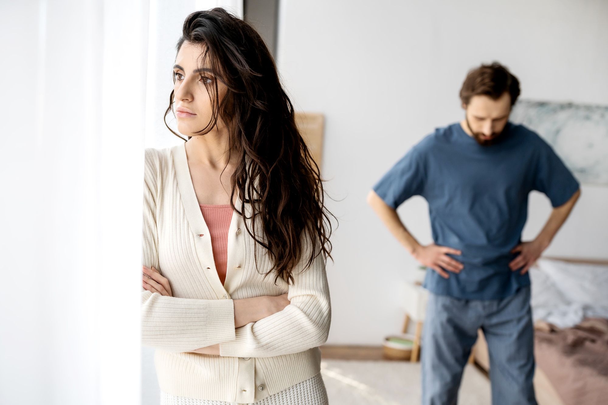 A woman with long dark hair stands by a window with her arms crossed, looking pensive and away from a man in the background who has his hands on his hips, appearing frustrated. The setting looks like a bedroom with soft lighting.