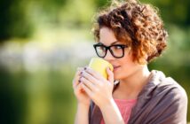 A person with curly hair and glasses is holding a yellow mug close to their mouth. They are wearing a pink top and a gray wrap, and they are outdoors with a blurred background of greenery. They appear to be enjoying a beverage.