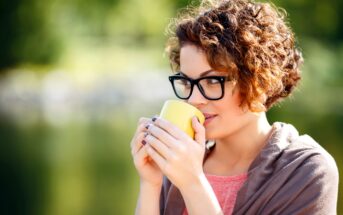 A person with curly hair and glasses is holding a yellow mug close to their mouth. They are wearing a pink top and a gray wrap, and they are outdoors with a blurred background of greenery. They appear to be enjoying a beverage.