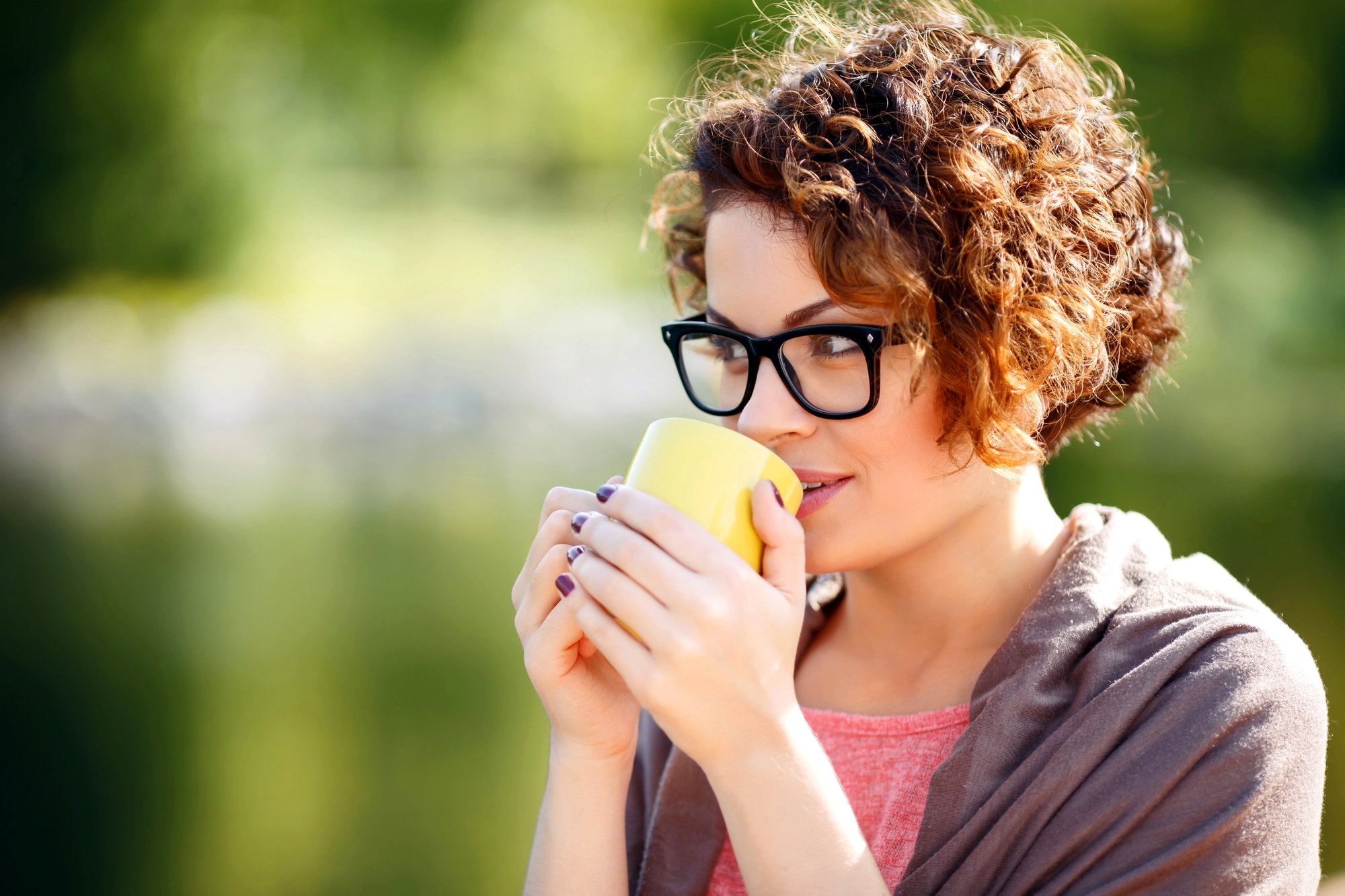 A person with curly hair and glasses is holding a yellow mug close to their mouth. They are wearing a pink top and a gray wrap, and they are outdoors with a blurred background of greenery. They appear to be enjoying a beverage.