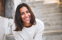 A young woman with shoulder-length brown hair wearing a white sweater is smiling brightly. She is leaning slightly forward and is outdoors, with a staircase and a partially visible stone wall in the background.