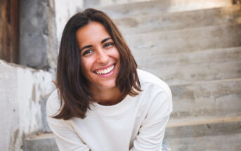 A young woman with shoulder-length brown hair wearing a white sweater is smiling brightly. She is leaning slightly forward and is outdoors, with a staircase and a partially visible stone wall in the background.