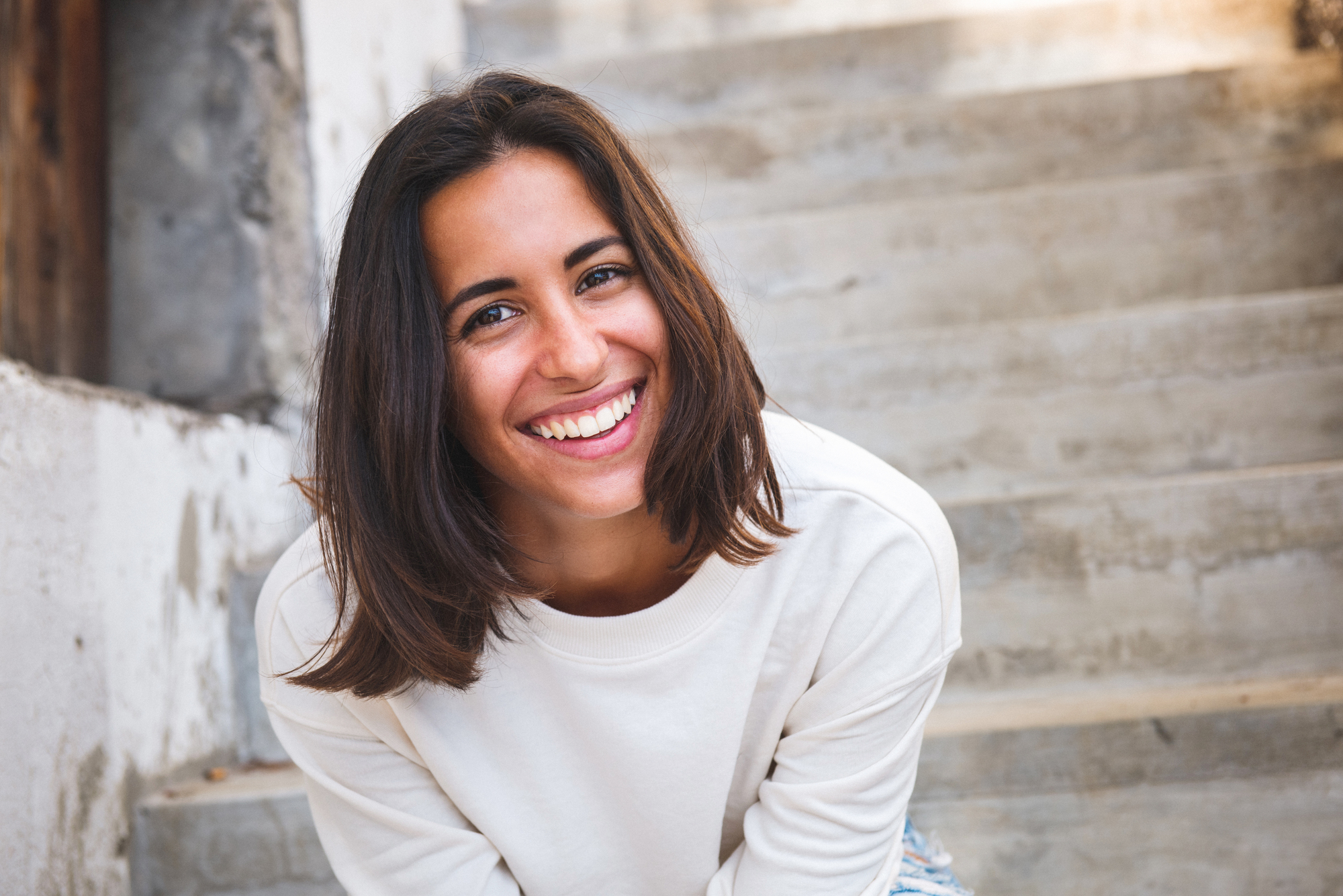 A young woman with shoulder-length brown hair wearing a white sweater is smiling brightly. She is leaning slightly forward and is outdoors, with a staircase and a partially visible stone wall in the background.