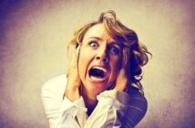 A woman with blond hair, wearing a white shirt, expresses intense fear or panic. Her eyes are wide open, and mouth agape. She holds her head with both hands against a muted background.