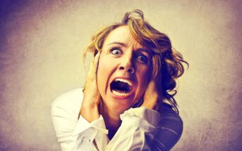 A woman with blond hair, wearing a white shirt, expresses intense fear or panic. Her eyes are wide open, and mouth agape. She holds her head with both hands against a muted background.