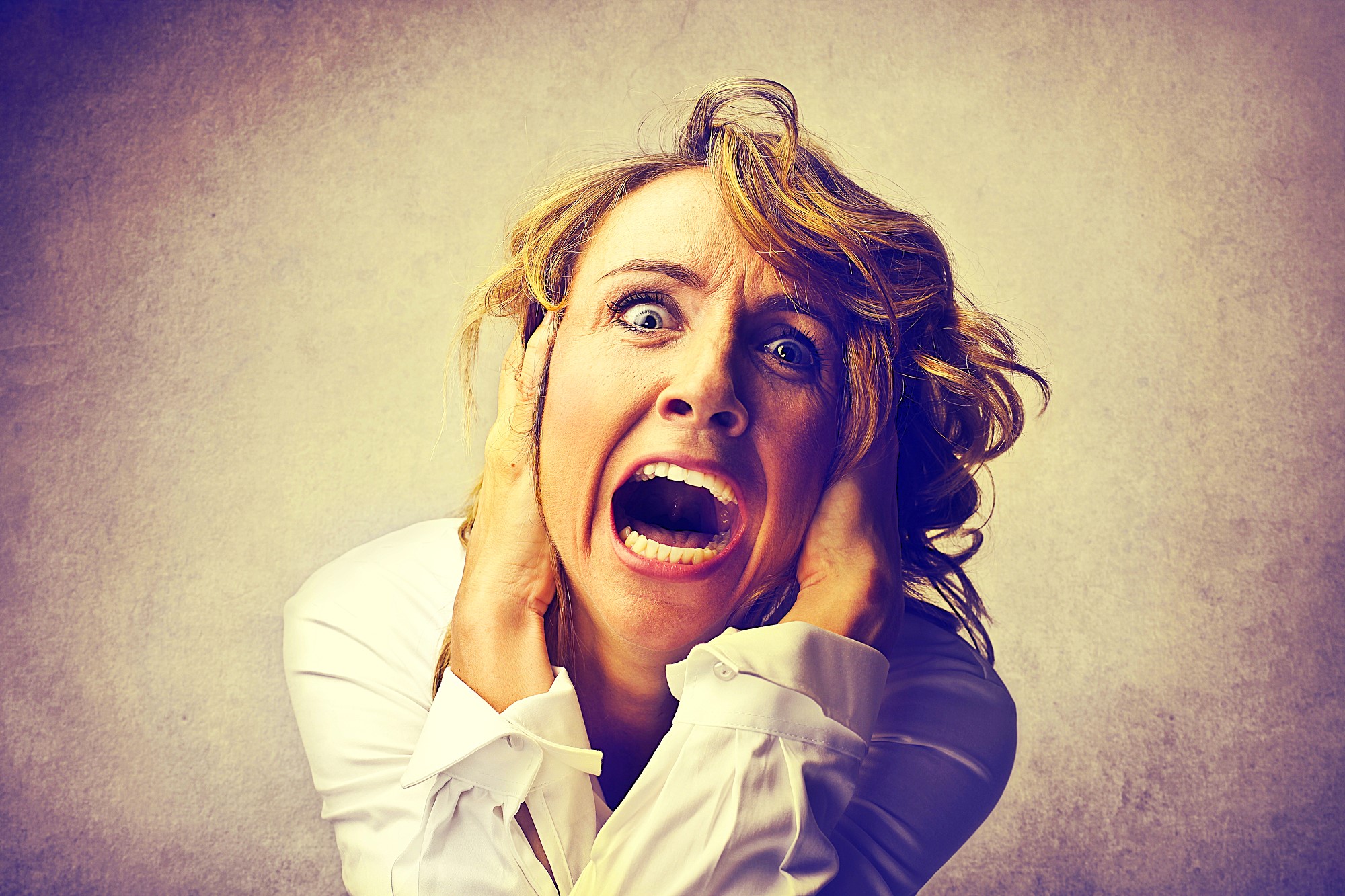 A woman with blond hair, wearing a white shirt, expresses intense fear or panic. Her eyes are wide open, and mouth agape. She holds her head with both hands against a muted background.