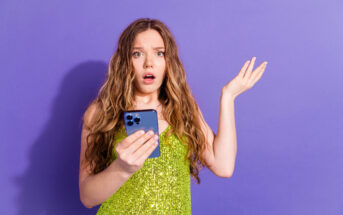 A young woman with long, wavy hair stands against a purple background, wearing a sparkly green dress. She holds a smartphone in one hand and has a surprised or confused expression, with her other hand raised in a questioning gesture.