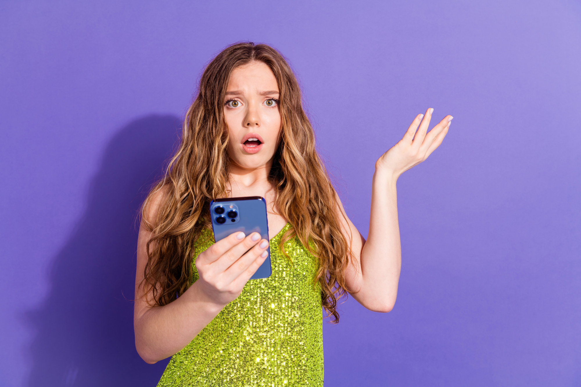 A young woman with long, wavy hair stands against a purple background, wearing a sparkly green dress. She holds a smartphone in one hand and has a surprised or confused expression, with her other hand raised in a questioning gesture.