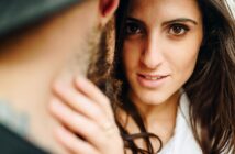 Close-up of a woman with long brown hair and deep brown eyes gazing intently at the camera. She is holding the face of a bearded man who is partially visible from the back, wearing a hat. The background is blurred, highlighting the subjects.