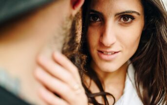 Close-up of a woman with long brown hair and deep brown eyes gazing intently at the camera. She is holding the face of a bearded man who is partially visible from the back, wearing a hat. The background is blurred, highlighting the subjects.