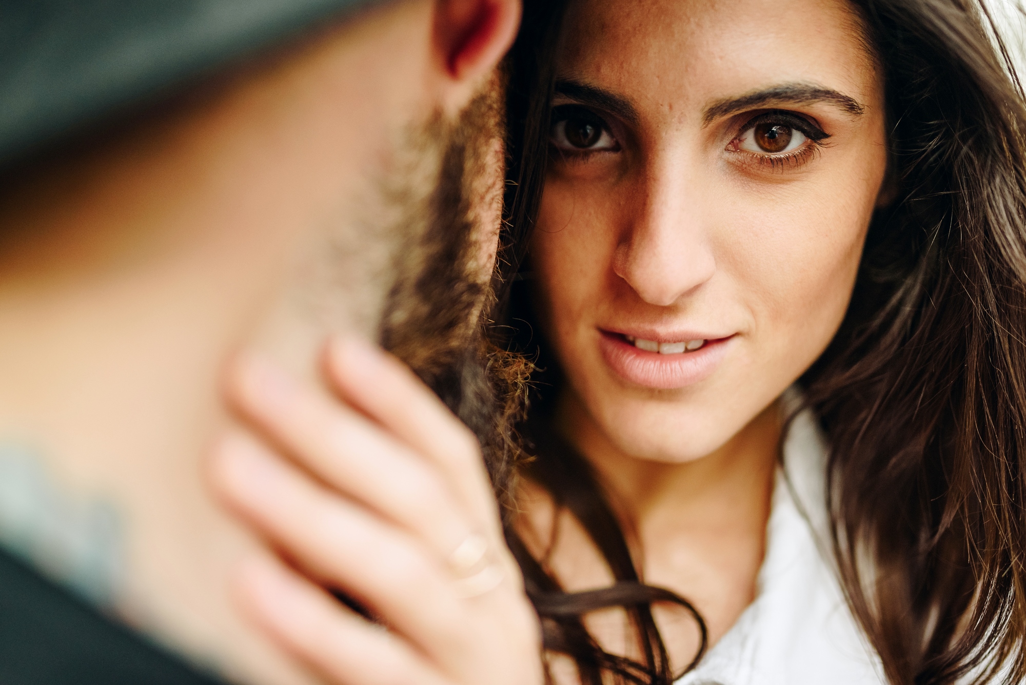 Close-up of a woman with long brown hair and deep brown eyes gazing intently at the camera. She is holding the face of a bearded man who is partially visible from the back, wearing a hat. The background is blurred, highlighting the subjects.