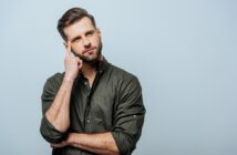 A bearded man in a dark green button-up shirt stands against a light blue background. He has one hand crossed against his chest and the other hand touching his temple, with a thoughtful expression on his face.