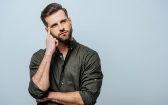 A bearded man in a dark green button-up shirt stands against a light blue background. He has one hand crossed against his chest and the other hand touching his temple, with a thoughtful expression on his face.