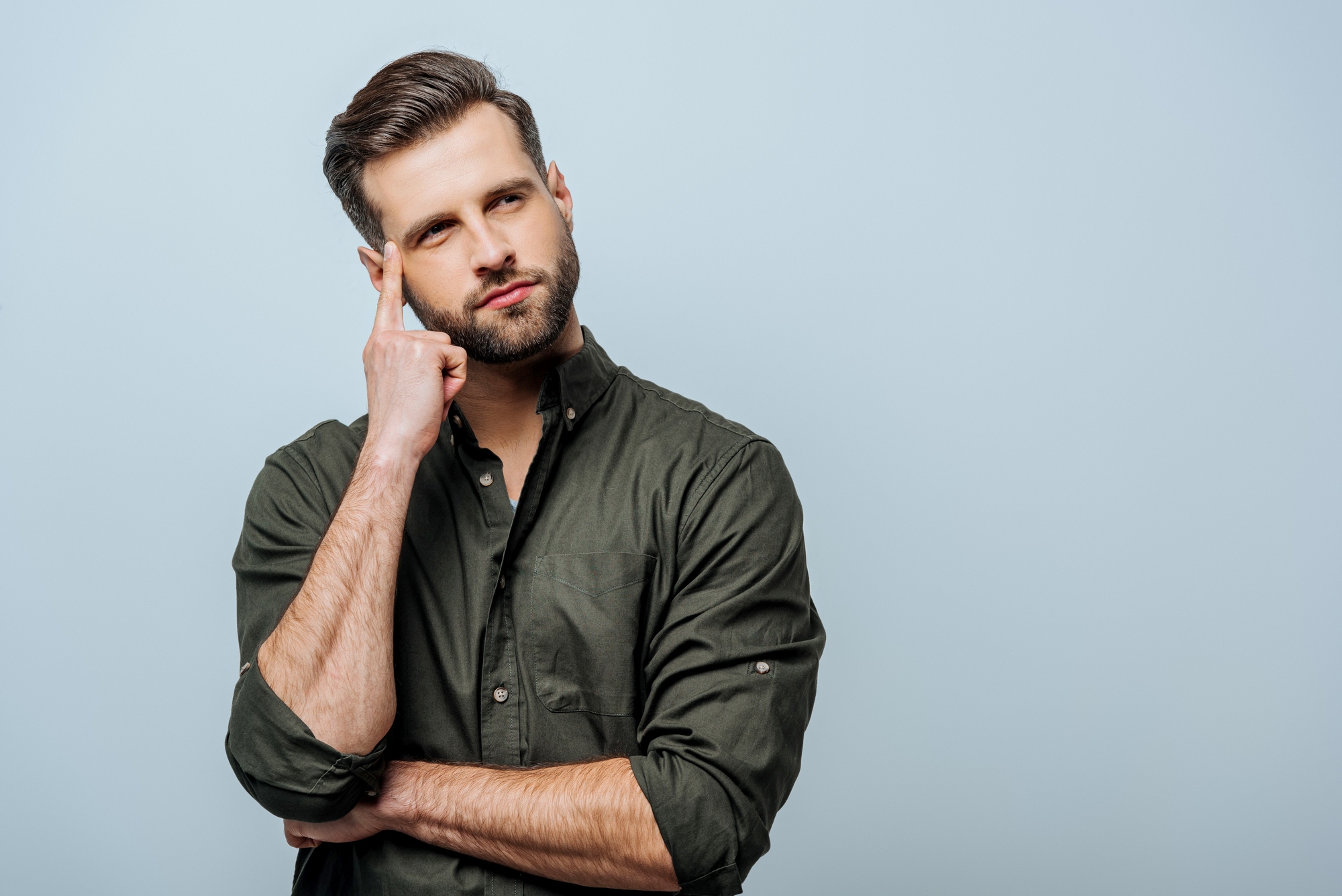 A bearded man in a dark green button-up shirt stands against a light blue background. He has one hand crossed against his chest and the other hand touching his temple, with a thoughtful expression on his face.