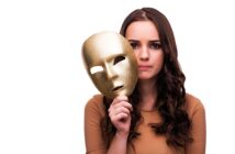 A woman with long, wavy brown hair holds a gold mask with empty eye holes in front of her face. She gazes at the camera with a neutral expression. She is wearing a beige top and has the mask positioned to the side of her face, revealing her features.