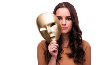 A woman with long, wavy brown hair holds a gold mask with empty eye holes in front of her face. She gazes at the camera with a neutral expression. She is wearing a beige top and has the mask positioned to the side of her face, revealing her features.