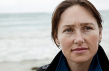 A woman with short brown hair stands near a beach, looking slightly to her right with a thoughtful expression. She wears a dark coat over a blue shirt, with the ocean in the background under a cloudy sky.