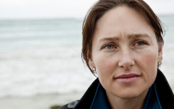 A woman with short brown hair stands near a beach, looking slightly to her right with a thoughtful expression. She wears a dark coat over a blue shirt, with the ocean in the background under a cloudy sky.