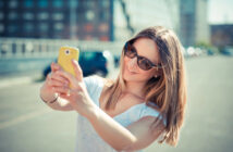 A woman with shoulder-length blonde hair, wearing sunglasses and a white T-shirt, is taking a selfie with her yellow smartphone on a sunny day. She is outdoors, and the background is slightly blurred, showing buildings and a few cars.