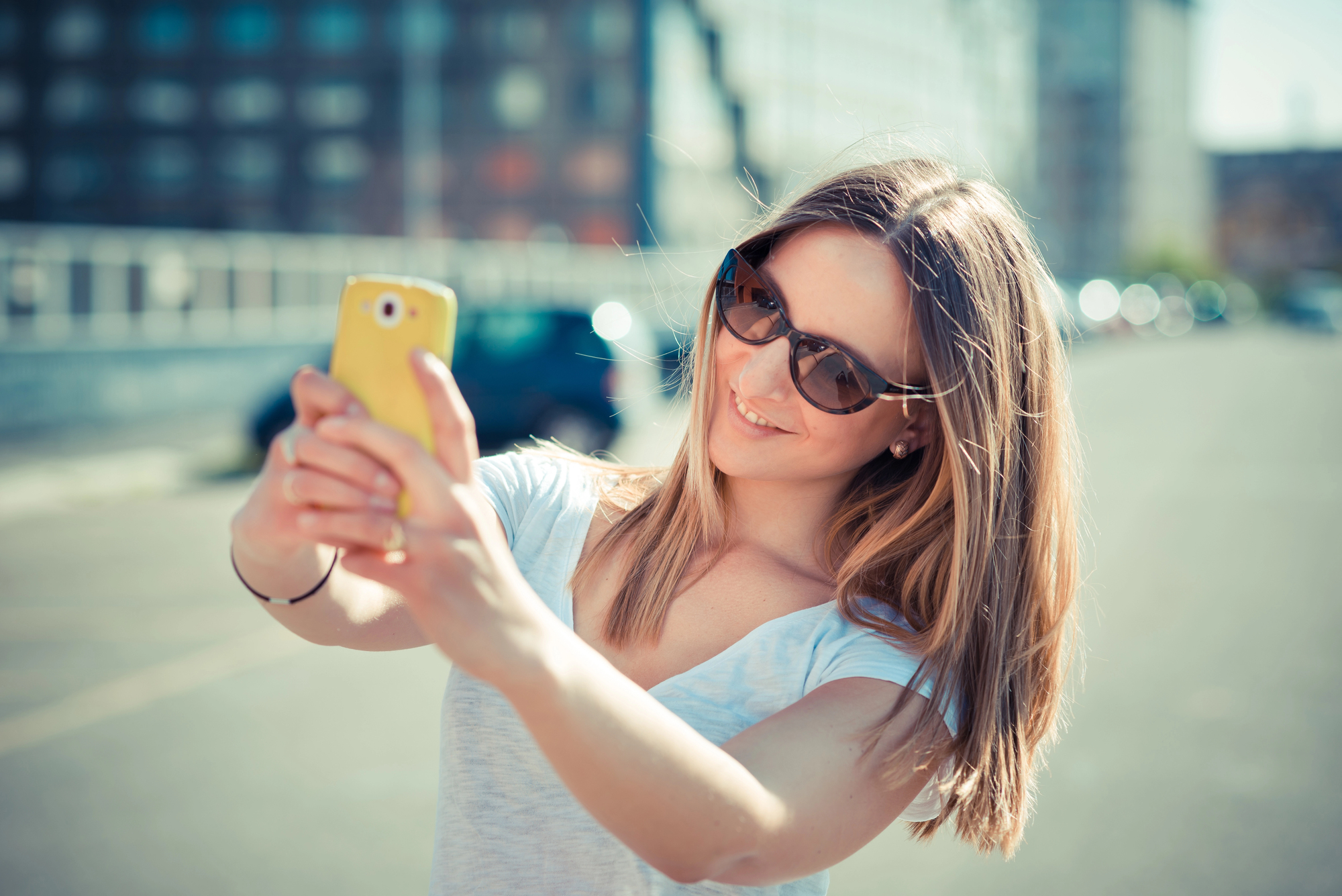 A woman with shoulder-length blonde hair, wearing sunglasses and a white T-shirt, is taking a selfie with her yellow smartphone on a sunny day. She is outdoors, and the background is slightly blurred, showing buildings and a few cars.