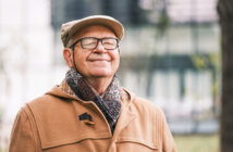 A smiling elderly man is outdoors, wearing a beige coat, a matching flat cap, and a cozy scarf. He has glasses and appears content as he stands in a blurred outdoor setting, possibly a park or garden area.